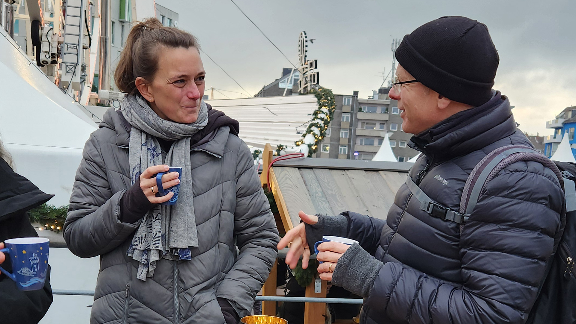 Links eine weiblich gelesene Person mit braunem Haar, welches zu einem Pferdeschwanz gebunden wurde. Sie trägt eine graue Jacke und grauen Schal und hält eine Tasse in der Hand. Sie blickt zu der männlich gelesenen Person rechts, welcher gerade etwas lustiges erzählt. Er trägt eine dunkle Mütze und eine schwarze Jacke.
