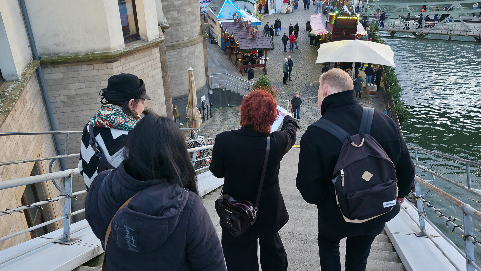 4 Menschen gehen eine Treppe herunter. Da sie von hinten fotografiert wurden, erkennt man ihre Gesichter nicht, sie tragen jedoch alle Winterjacken und dunkle Kleidung. Die Person in der Mitte zeigt mit deren Zeigefinger die Richtung an, in die sie gehen wollen. Im Hintergrund sehen wir ein Riesenrad.