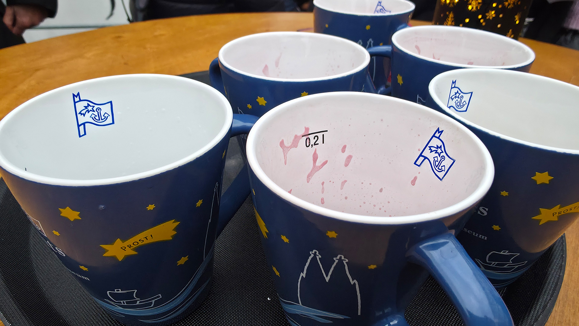 Sechs blaue Keramiktassen mit maritimem Design stehen auf einem Tablett auf einem Holztisch. Einige enthalten Reste eines pinken Getränks, der Schriftzug „PROST!“ ist gut sichtbar.