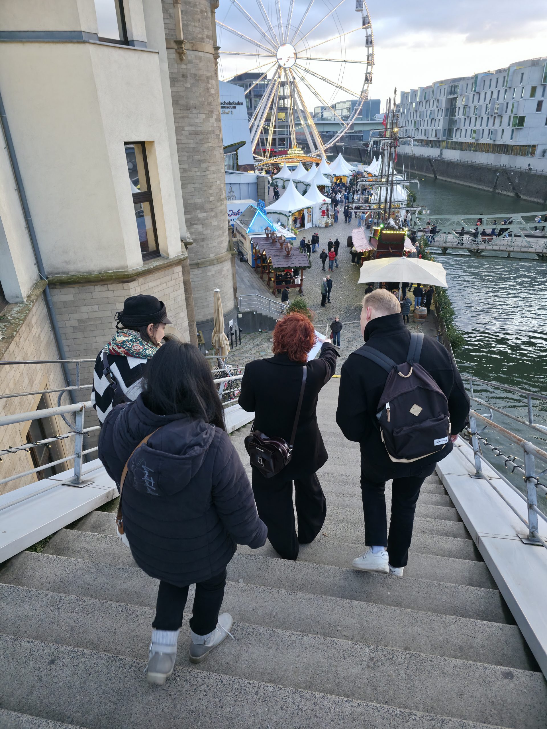 4 Menschen gehen eine Treppe herunter. Da sie von hinten fotografiert wurden, erkennt man ihre Gesichter nicht, sie tragen jedoch alle Winterjacken und dunkle Kleidung. Die Person in der Mitte zeigt mit deren Zeigefinger die Richtung an, in die sie gehen wollen. Im Hintergrund sehen wir ein Riesenrad.