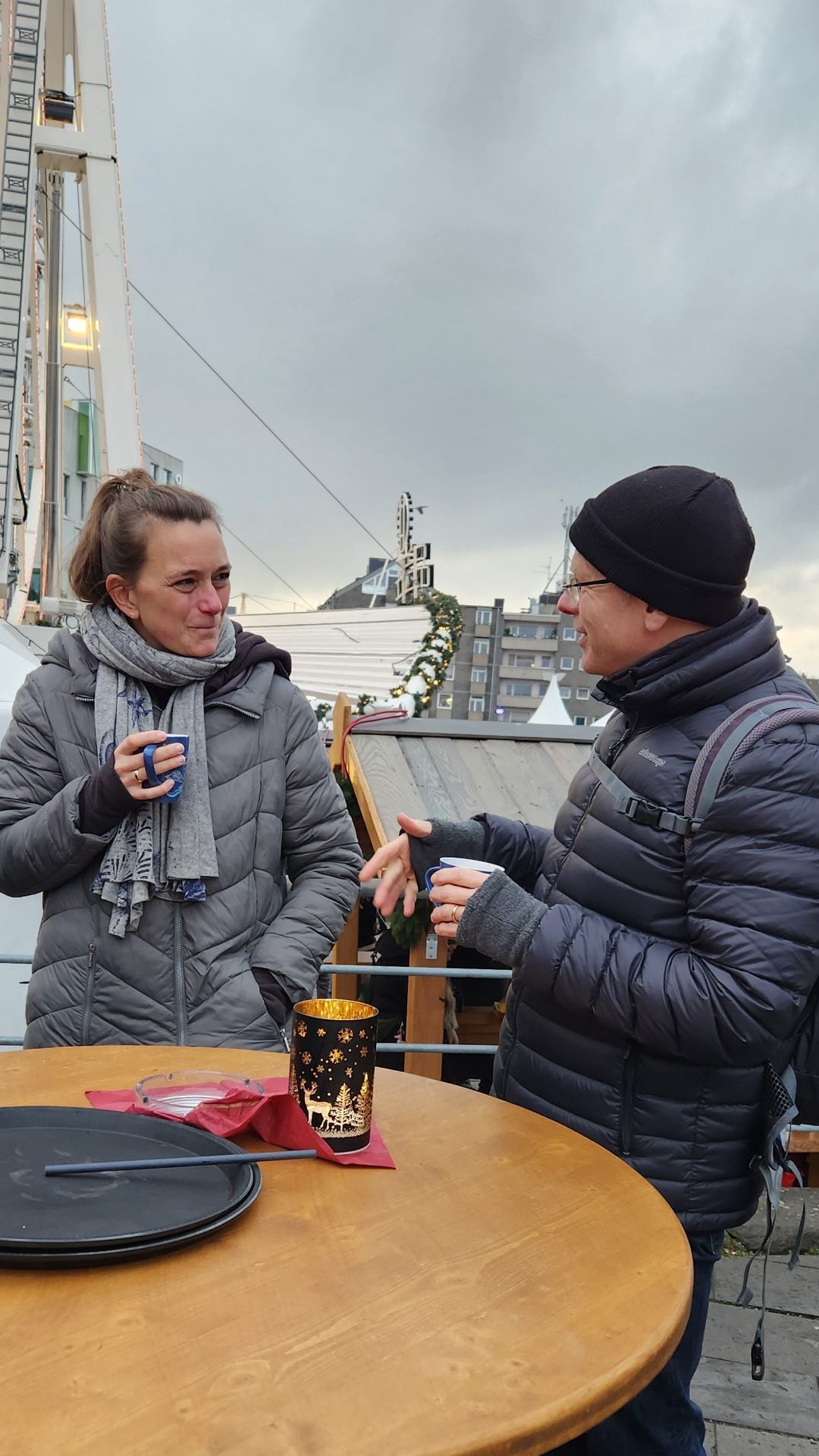 Links eine weiblich gelesene Person mit braunem Haar, welches zu einem Pferdeschwanz gebunden wurde. Sie trägt eine graue Jacke und grauen Schal und hält eine Tasse in der Hand. Sie blickt zu der männlich gelesenen Person rechts, welcher gerade etwas lustiges erzählt. Er trägt eine dunkle Mütze und eine schwarze Jacke.