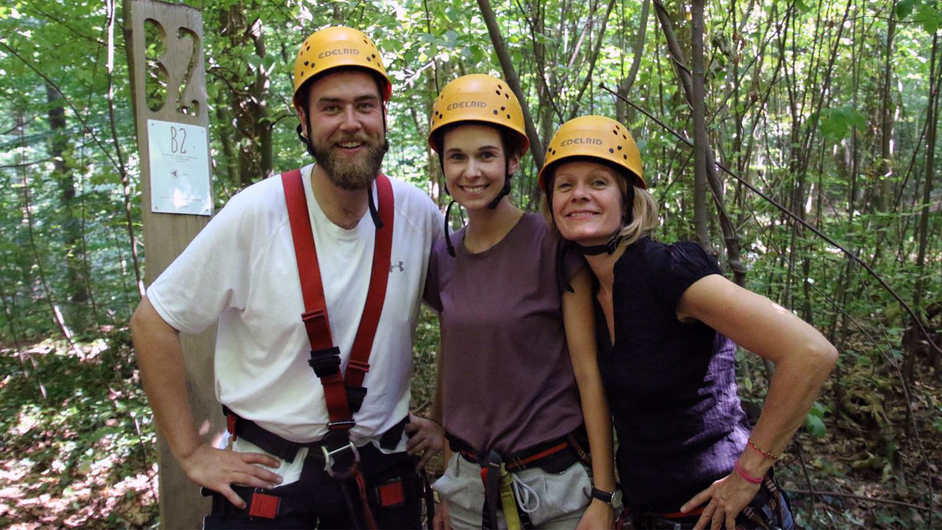 Drei Personen stehen eng beieinander in einem bewaldeten Außenbereich. Sie tragen gelbe Schutzhelme und Klettergurte, was auf eine Aktivität wie Hochseilgarten, Kletterparcours oder Ziplining hinweist. Hinter ihnen ist dichtes grünes Grün aus Bäumen und Sträuchern zu sehen. Links im Bild steht ein Holzpfosten mit der Markierung „B2“ und einem kleinen Schild darunter, das ebenfalls „B2“ zeigt. Die Szene vermittelt den Eindruck eines gemeinsamen Outdoor‑Erlebnisses, bei dem Sicherheit und Teamgefühl im Vordergrund stehen.