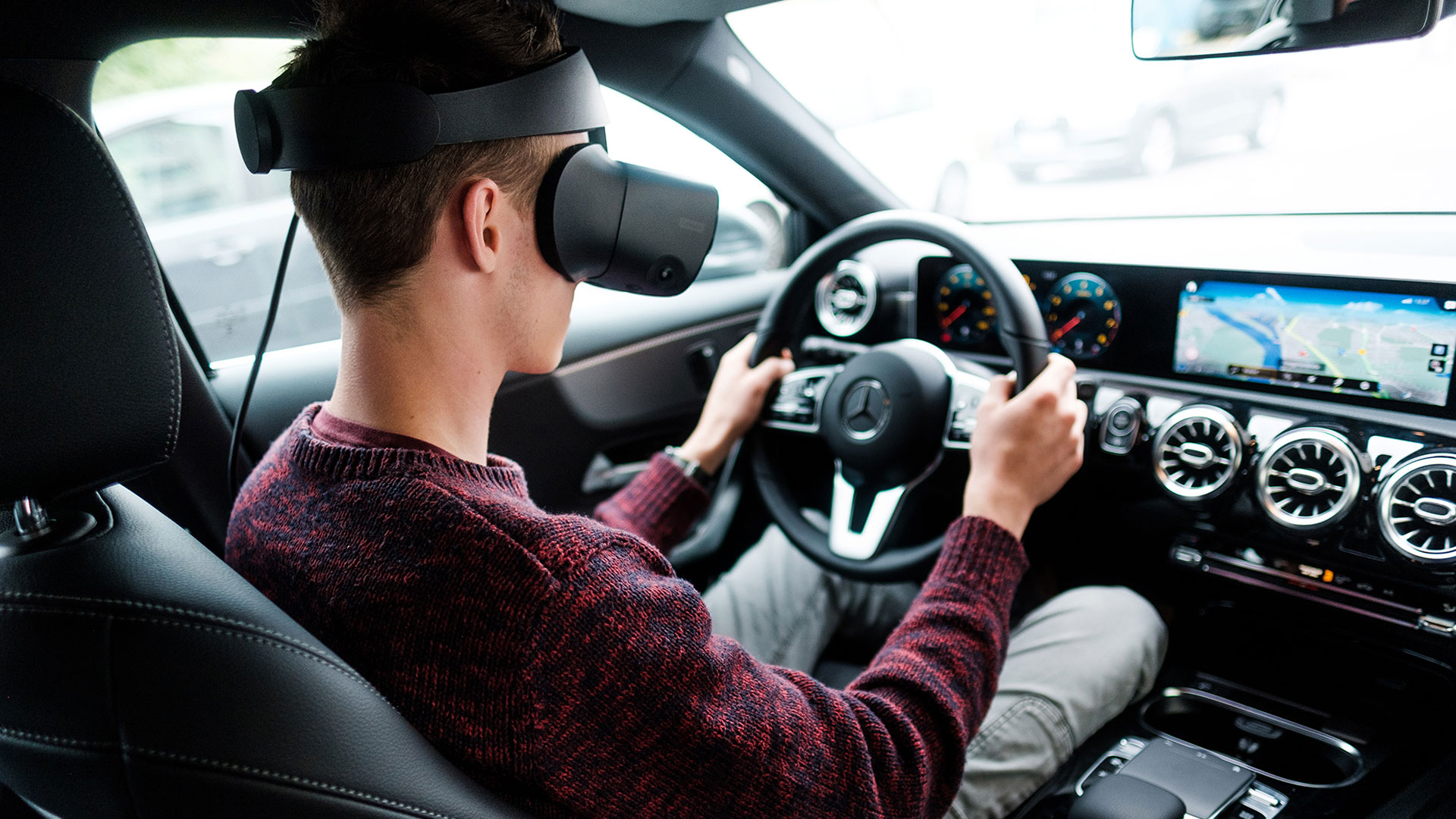 Person sitzt im Fahrersitz eines modernen Mercedes‑Benz, trägt ein VR‑Headset und hält das Lenkrad; digitales Cockpit und großes Infotainment‑Display mit Navigationskarte im Hintergrund.