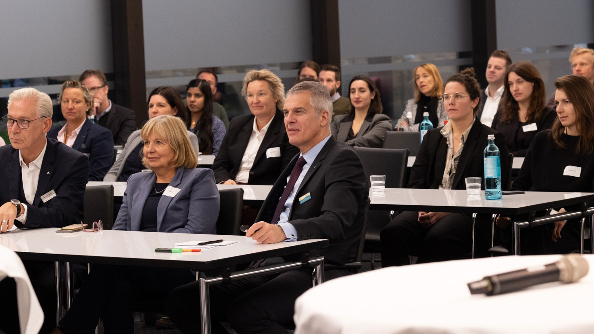 Mehrere Personen sitzen in einem Konferenz‑ oder Seminarraum und hören aufmerksam einer Präsentation oder einem Vortrag zu. Die Teilnehmenden tragen überwiegend Business‑Kleidung, vor ihnen liegen Notizbücher, Stifte und Wasserflaschen. Im Vordergrund steht ein Mikrofon auf einem Tisch, was darauf hinweist, dass eine Sprecherin oder ein Sprecher gleich spricht oder bereits gesprochen hat. Der Raum wirkt hell und professionell, mit großen Fenstern und teilweise heruntergelassenen Jalousien. Die Atmosphäre vermittelt den Eindruck eines formellen Meetings, Workshops oder einer geschäftlichen Veranstaltung.