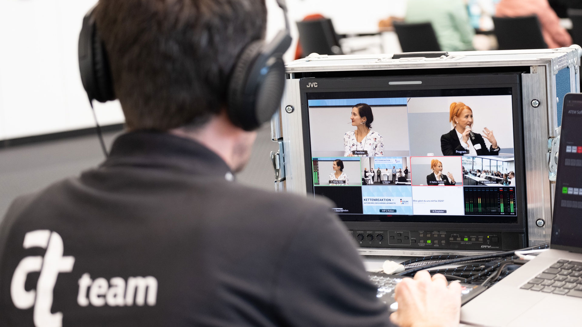 Eine Person sitzt vor einem professionellen Videoproduktions‑Setup und trägt Kopfhörer sowie ein schwarzes Shirt mit der Aufschrift „ateam“ auf dem Rücken. Vor ihr steht ein Monitor, der mehrere Video‑Feeds eines laufenden Events zeigt – darunter verschiedene Kameraeinstellungen von Sprecher*innen und Präsentationsfolien. Die Benutzeroberfläche zeigt typische Elemente einer Live‑Regie wie „Preview“, „Program“ und Audiopegelanzeigen. Die Szene vermittelt den Eindruck einer konzentrierten technischen Arbeit hinter den Kulissen einer Konferenz oder Live‑Übertragung, bei der mehrere Kameras und Tonquellen koordiniert werden.