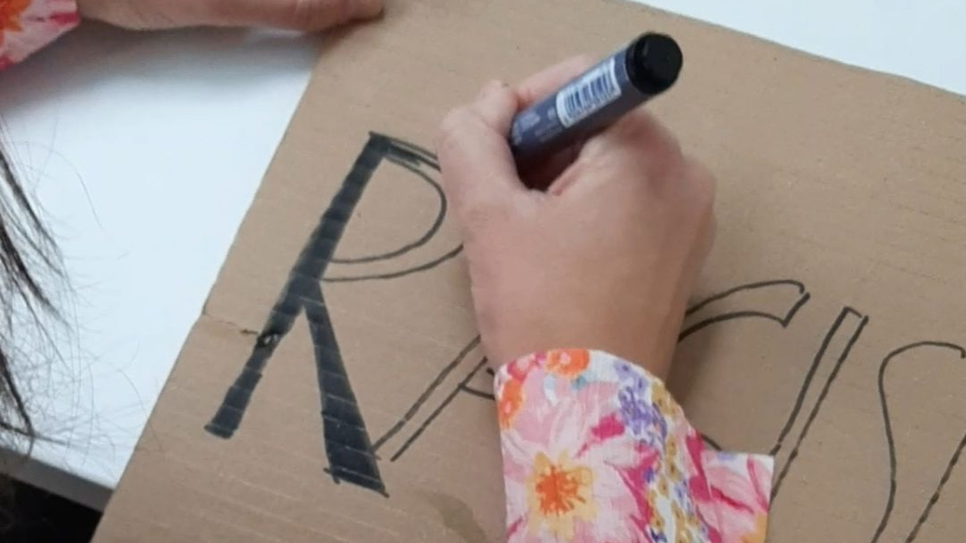 Eine Person schreibt mit einem schwarzen Marker auf ein Stück Karton. Der Karton liegt auf einer glatten Oberfläche, und das Wort beginnt mit den Buchstaben „RACISM“, was darauf hinweist, dass ein Statement oder Plakat entsteht. Die Person trägt ein farbenfrohes, floral gemustertes Oberteil in Pink‑, Orange‑ und Lilatönen.