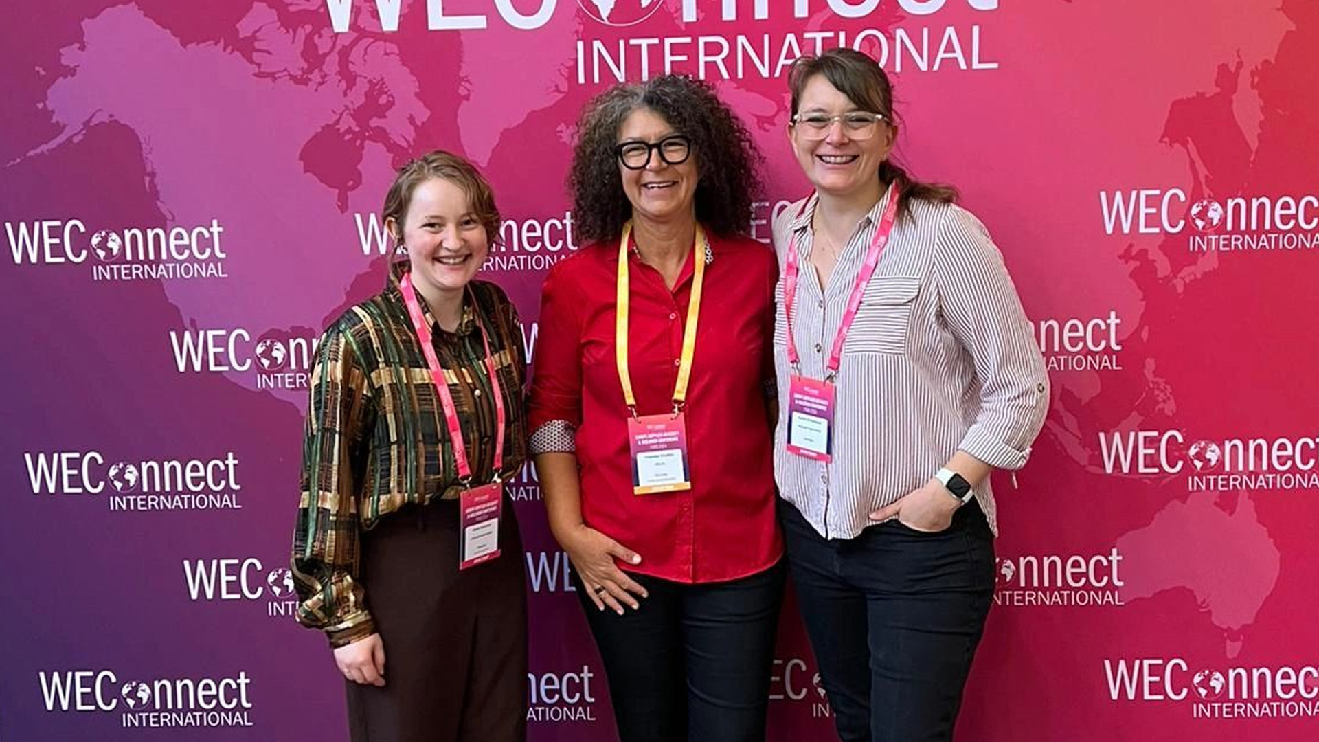 Drei Personen stehen vor einem pink‑violetten Hintergrund mit dem mehrfach wiederholten Schriftzug „WEConnect International“. Der Hintergrund zeigt eine stilisierte Weltkarte. Alle drei tragen Namensschilder an Lanyards, was auf eine Konferenz oder ein Networking‑Event hinweist. Die Personen stehen nebeneinander, lächeln in die Kamera und wirken wie Teilnehmende oder Vertreter einer internationalen Business‑Veranstaltung.